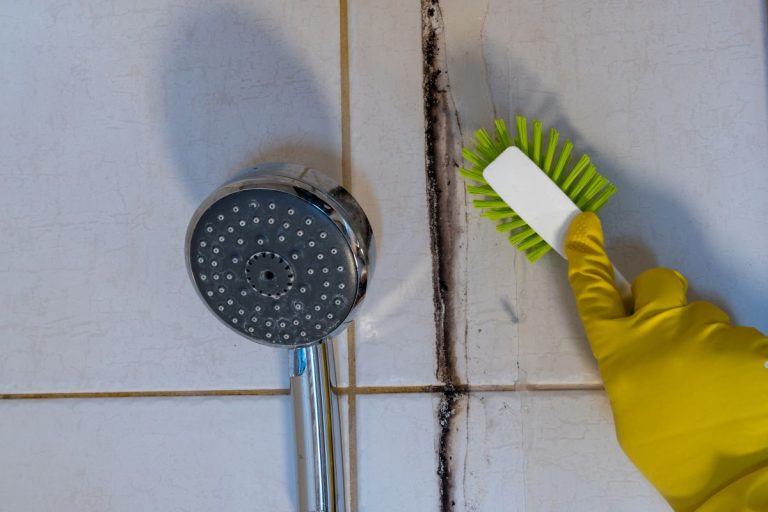 A person wearing a yellow glove cleans a shower head, focusing on removing mold and ensuring a hygienic bathroom.
