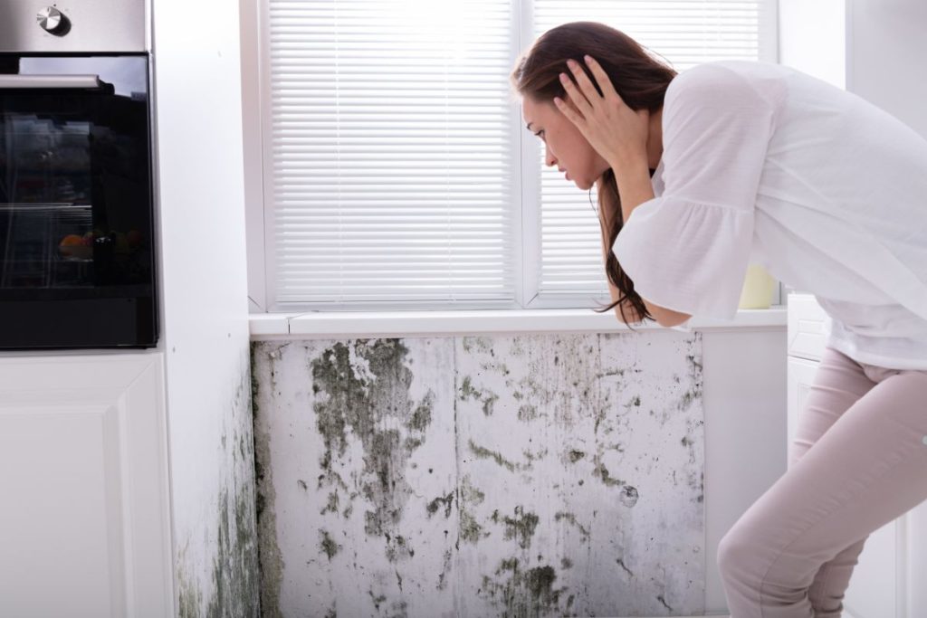 A woman stands before a moldy wall, highlighting the health risks of inhaling mold spores, including cancer.